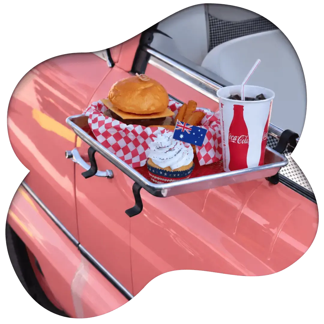 A curbside food tray mounted on a car window holding a burger, fries, a cupcake, and a drink for pickup